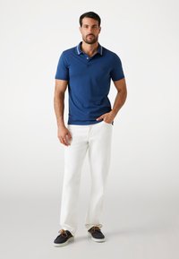 Polo shirt blu con colletto a contrasto, abbinato a pantaloni bianchi e scarpe scure. Presenta un piccolo logo sul lato sinistro del petto.
