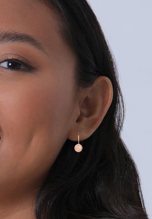 Orecchini a cerchio in oro rosa con un piccolo charm circolare e texturizzato appeso a un sottile e semplice gancio. Inquadratura ravvicinata su uno sfondo neutro.