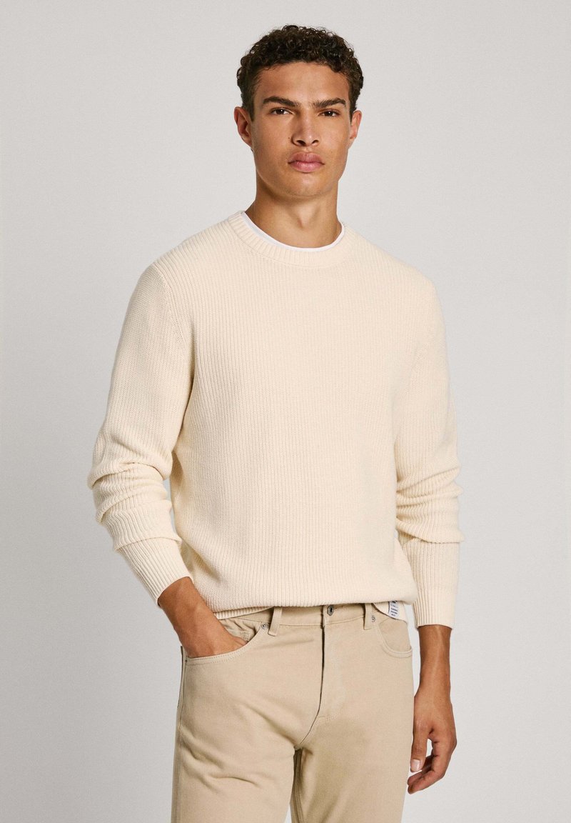 Bež pulover iz rebraste pletenine s krojnim ovratnikom in dolgimi rokavi, v kombinaciji z svetlobeznimi hlačami. Teksture tkanine z sproščenim krojem.