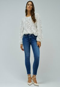 Blusa blanca con volantes y mangas largas, combinada con jeans ajustados azules y tacones blancos de punta, sobre un fondo simple.