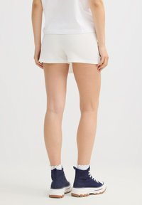Mujer que lleva pantalones cortos blancos con textura, top blanco, calcetines blancos con volantes y zapatillas altas de color azul marino, de pie sobre un fondo liso.