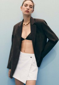 Blazer noir sur un haut de bikini noir, associé à une mini jupe enveloppante blanche avec un détail de bouton noir. Tissu lisse et design structuré.