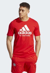 La camiseta roja de Adidas del FC Bayern presenta un logo blanco y tres franjas. Combinada con pantalones cortos rojos a juego, exhibe un look atlético clásico.