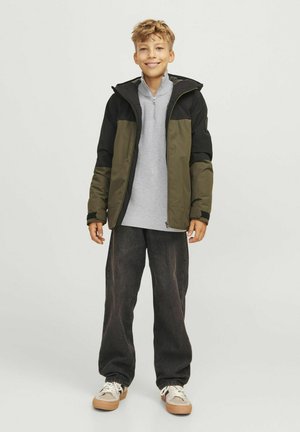 Lättvikts, zipprad huva jacka i olivgrön och svart, tillsammans med en grå långärmad tröja, mörka löst sittande byxor och beige sneakers.