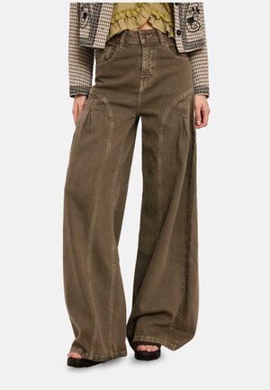 Pantalones marrones de pierna ancha con costuras detalladas, combinados con sandalias negras abiertas, un top verde con volantes y una chaqueta estampada.