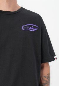 Black cotton t-shirt featuring a purple "REplay" logo on the chest. Short sleeves with a tag visible on the hem. Smooth texture.