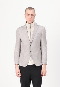 Blazer gris clair avec une finition texturée, doté de deux poches avant et d'une fermeture à boutons, superposé à un vêtement crémeux à col haut.