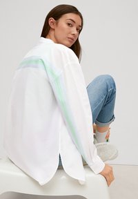 White long-sleeve shirt with light green and blue stripes, slight high-low hem, worn with rolled-up blue jeans and light gray sneakers.