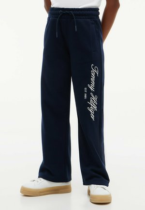 Person iført marineblå joggingbukser med "Tommy Hilfiger EST 1985" trykt på venstre ben og hvide sneakers med gummisåler.