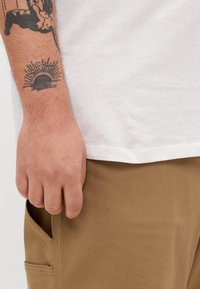 Hvid bomulds t-shirt med korte ærmer, båret med tanfarvede bukser med en sidelomme. Tatovering på armen viser et sol-design.