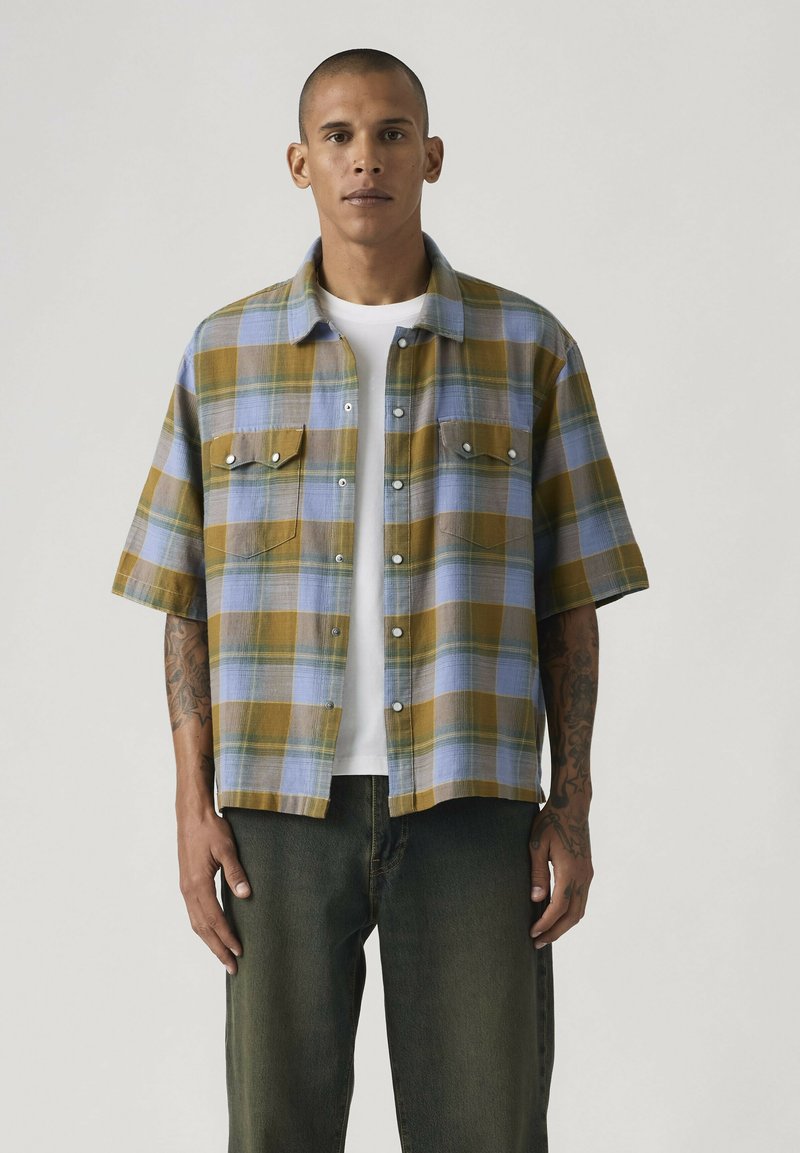 Man draagt een kortmouws geruit overhemd in blauw en mosterdkleur over een wit T-shirt, gecombineerd met donkergroene broek, staand tegen een effen achtergrond.