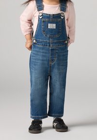 Modré džínové lacláče s nastavitelnými popruhy, přední kapsou a širokými nohavicemi. Doplněné černými teniskami. Bavlněná látka, ležérní vzhled.