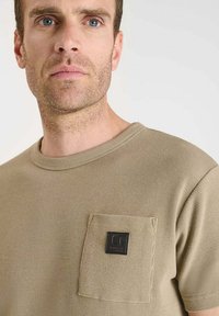 Camisa de manga corta beige con una tela texturada de punto gofrado y un bolsillo de parche, con una etiqueta de logo negra en el lado izquierdo.