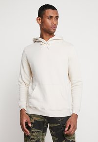 Beige genser laget av myk stoff, med snorhette, kengurulomme og ribbestrikkede mansjetter, kombinert med kamuflasjefargede cargo-bukser.
