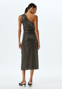 Asymmetrisches schwarzes Kleid mit schimmernder Textur, knielang, figurbetonter Silhouette, mit einem Rückendrehdetail, ergänzt durch Slingback-Absätze.