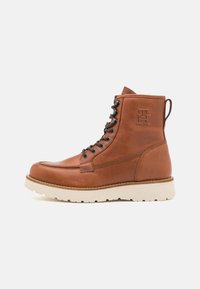 Brown leather boot with lace-up design, round toe, and a white rubber sole. Features embossed logo and contrasting stitching details.