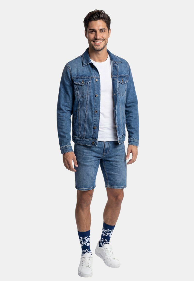 Mann lächelnd, trägt blaue Jeansjacke und Shorts, weißes T-Shirt, gemusterte blaue Socken und weiße Sneakers, steht vor weißem Hintergrund.