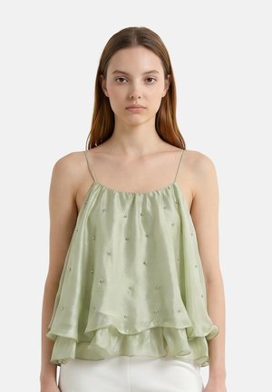 Giovane donna con capelli castani lisci che indossa un top verde chiaro senza maniche a strati con sottili spalline e piccole decorazioni con perline.