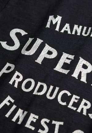 Schwarzer Stoff mit weißen, rissigen Buchstaben. Der Text enthält "Manuf" und "Super" sowie "Producers". Der Fokus liegt auf einem Vintage-Stil und einer strukturierten Oberfläche.