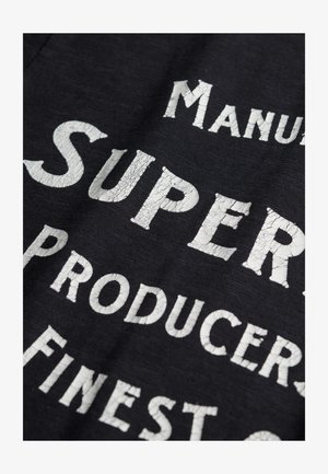 Tissu noir avec des lettres blanches fissurées. Le texte inclut "Manuf" et "Super" accompagnés de "Producers". Accent sur le style vintage et la surface texturée.