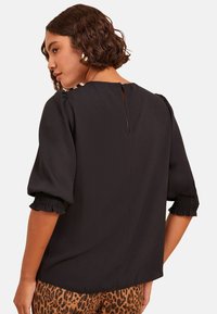 Donna con capelli ricci e corti, che indossa un top nero con maniche a sbuffo e polsini arricciati, e pantaloni con stampa leopardata, in profilo davanti a uno sfondo bianco.