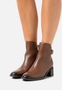 Botines de cuero marrón con un pequeño tacón cuadrado. Cuentan con una cremallera lateral y un acento decorativo de hebilla. Textura suave con un acabado pulido.