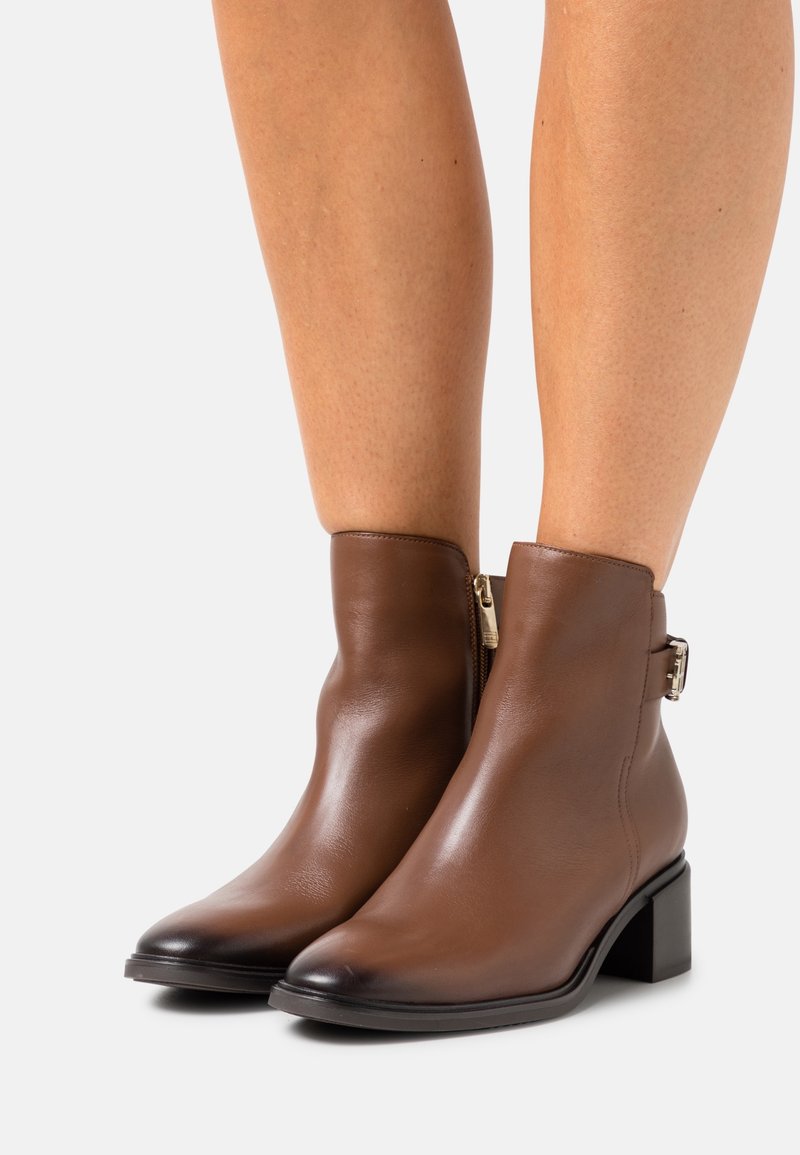 Botines de cuero marrón con un pequeño tacón cuadrado. Cuentan con una cremallera lateral y un acento decorativo de hebilla. Textura suave con un acabado pulido.