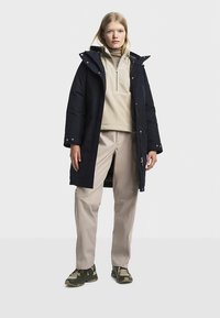 Navy blue waterproof parka with a hood, tan fleece top, and beige pants. Features buttons and zippers, layered for warmth and functionality.