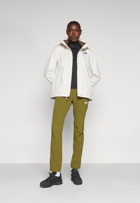 Giacca impermeabile bianca con una fodera interna marrone, top nero e pantaloni verde oliva. Scarpe da trekking nere completano l'outfit.