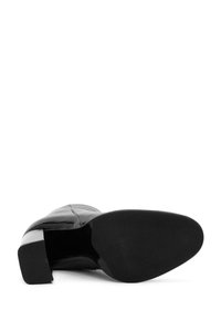 Botte de cheville en cuir noir avec un bout carré, un talon quadratique et une surface lisse. La semelle est plate et texturée pour une bonne adhérence, contrastant avec le dessus brillant.