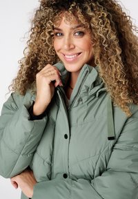 Gevoerde groene jas met een rits aan de voorkant, zwarte drukknoopsluitingen en een verstelbare capuchon; gemaakt van glad, geplooid materiaal met een zachte afwerking.