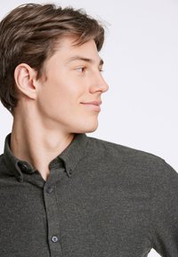 Camisa de botón verde oscura hecha de tela texturizada, con cuello, botones negros y un logo sutil sobre el pecho izquierdo.