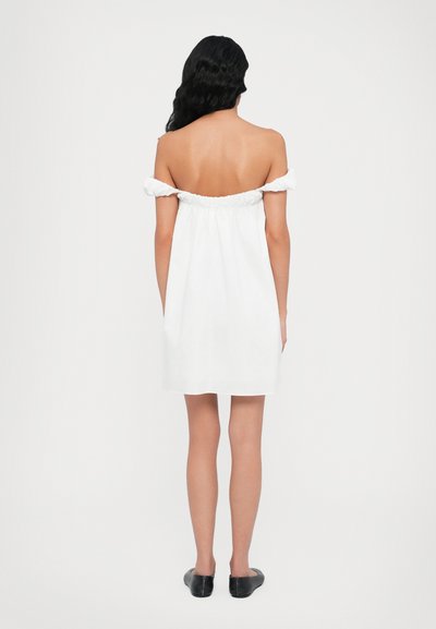 Rochie albă cu umeri goi, cu guler plisat. Material neted, croială lejeră, lungime până la mijlocul coapsei, asortată cu pantofi negri tip slip-on.