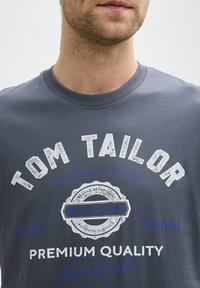 Camiseta de algodón gris con cuello redondo, impresión desgastada en blanco y azul de "Tom Tailor" y el texto "Calidad Premium", con detalle de logo circular.