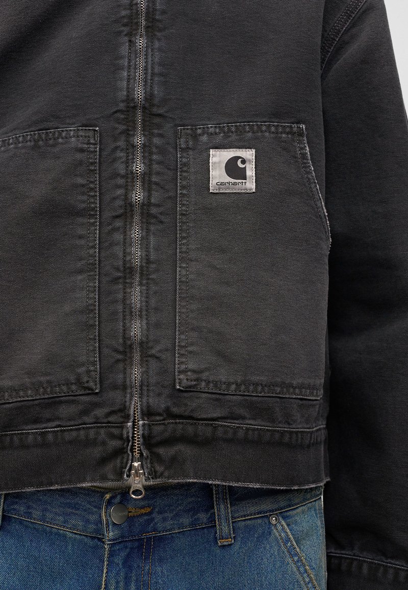 Veste noire Carhartt zippée avec poches avant portée sur un jean en denim bleu, montrant la fermeture éclair et le patch logo de la marque sur la poche gauche.
