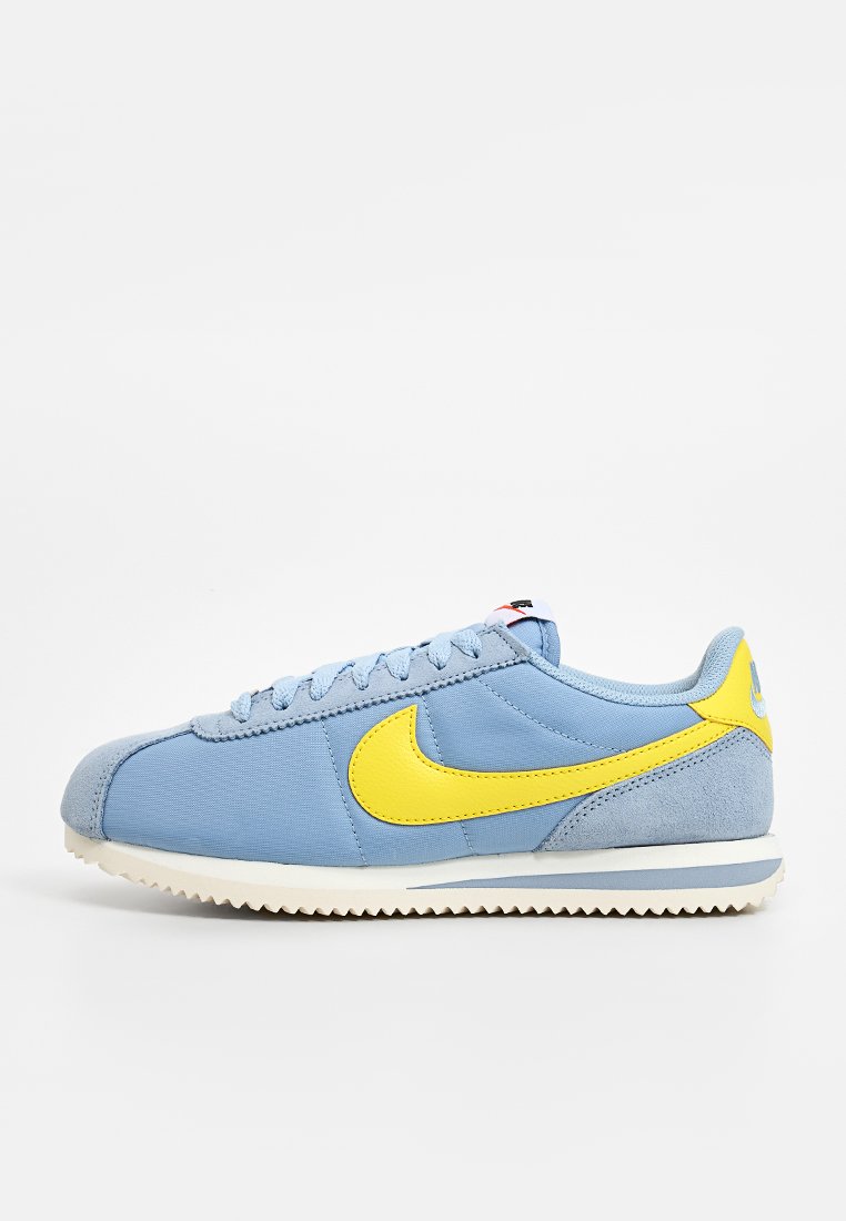 Baskets de sport bleu clair avec un swoosh jaune et des accents, présentant un talon en cuir suédé texturé, une semelle blanche en caoutchouc plate et un design à lacets.