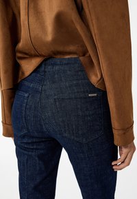 Donkerblauwe jeans met een aansluitend ontwerp, voorzien van twee achterzakken en subtiele stikseldetails, gecombineerd met een bruine, gestructureerde blouse.