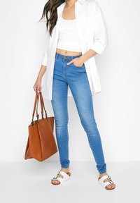 Weißes Hemd mit Knopfleiste über einem weißen Croptop, blaue Skinny-Jeans, braune Tragetasche und weiße Sandalen mit Schnallen.