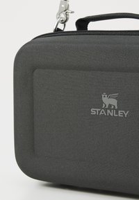 Musta kangaskotelo, jossa on teksturoidun pinnan, vetoketjullinen sulkeminen ja metallinen klipsi. Etupuolella on kohokuvioitu "STANLEY"-logo.