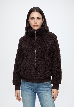Mujer con una chaqueta marrón oscura texturizada con cremallera y cuello alto, y vaqueros azules, de pie frente a un fondo blanco liso.