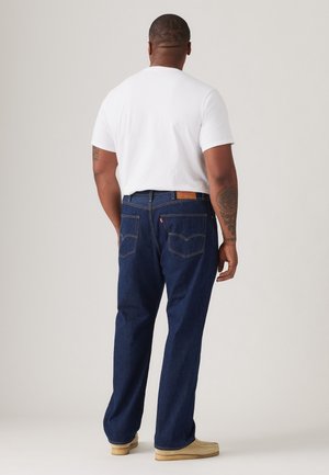 Mężczyzna w białej koszuli z krótkim rękawem, ciemnoniebieskich dżinsach, jasnobrązowych butach, zwrócony tyłem, pokazuje plecy i tatuaże na ramionach na jednolitym tle.