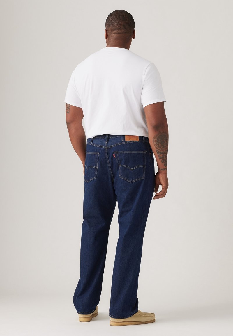 Homme portant une chemise blanche à manches courtes, un jean bleu foncé, des chaussures marron clair, de dos, montrant son dos et des tatouages sur les bras sur un fond uni.