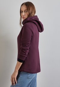 Felpa bordeaux con una texture morbida, dotata di un grande cappuccio, maniche aderenti e un design rilassato, abbinata a jeans azzurri chiari.