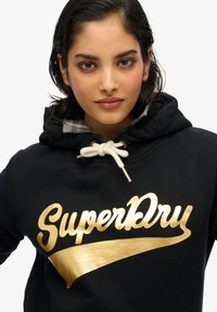 Sudadera negra con un logo y diseño dorado de "SuperDry", que presenta una capucha con cordón, forro de cuadros y material de algodón suave.
