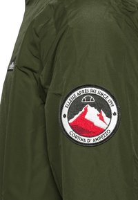 Giacca verde oliva con un patch ricamato circolare con montagne rosse e testo: “Ellesse Apres Ski Dal 1959 Cortina D'Ampezzo.”