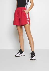 Red athletic shorts with white side stripes, elastic waistband, and pockets. Worn with black and white sneakers, featuring a logo on the side.