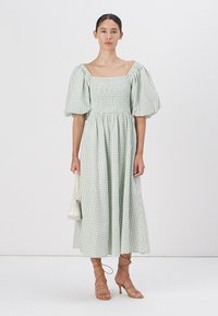 Šviesiai žalia gingham suknelė su pūstomis rankovėmis ir susiraukšlėtu liemeniu, turinti sruvenančią sijoną. Modelis laiko mažą kreminę rankinę ir dėvi batus su dirželiais.