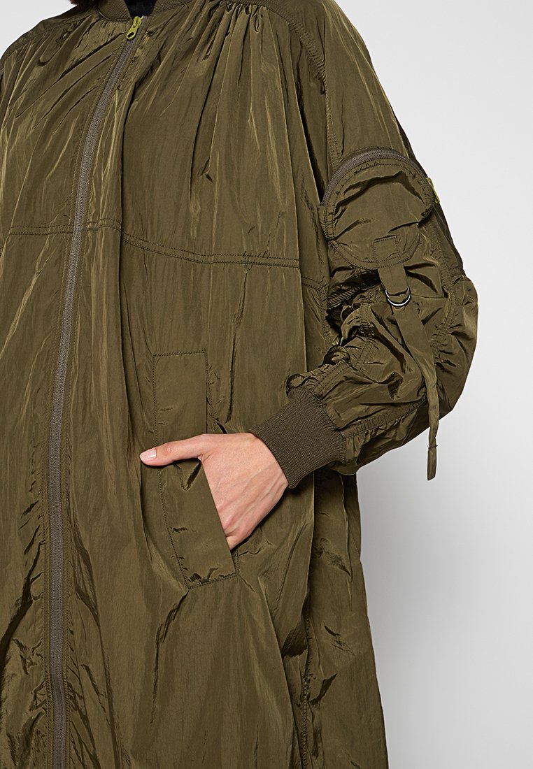 Jasnooliwkowy, lekki, długi płaszcz z przednim zamkiem, elastycznymi mankietami i kieszenią na rękę, z marszczonymi ramionami i regulowanymi detalami.