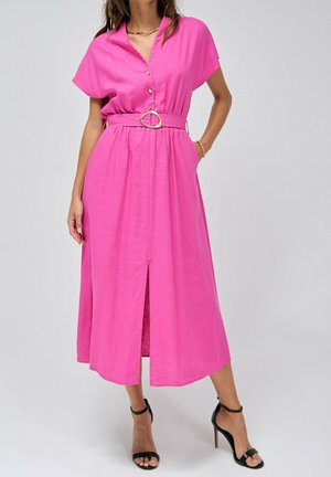 Femme portant une robe midi rose vif à manches courtes, ceinture à boucle dorée, et sandales à talons noirs, debout avec une main dans la poche.