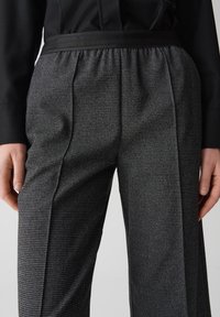 Grijze gemusterde broek met een getailleerde pasvorm. Heeft een zwarte tailleband, geplooide voorkant en een gladde textuur. Zakken zichtbaar aan de zijkanten.
